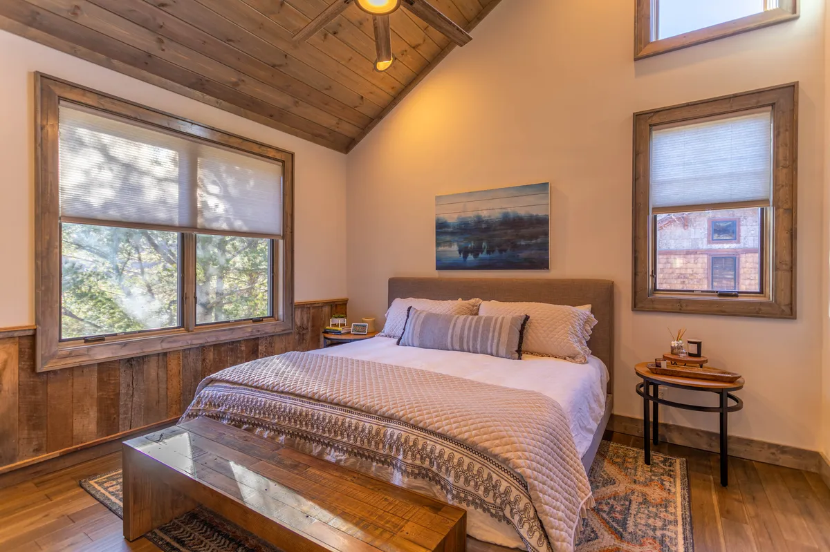 A serene bedroom with a king-sized bed adorned with a textured throw and pillows, wooden framed windows bringing in natural light, a tasteful art piece on the wall, and a wooden side table. The space exudes calm and comfort, perfect for restful retreats.