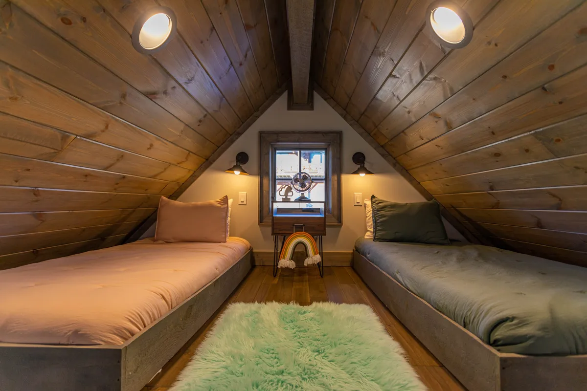 A playful and inviting loft space featuring two twin beds against a wall adorned with a decorative rainbow, soft bedding, and exposed wooden beams. A window allows natural light to brighten the cozy area, creating an ideal nook for children or friends to rest, read, or play games.