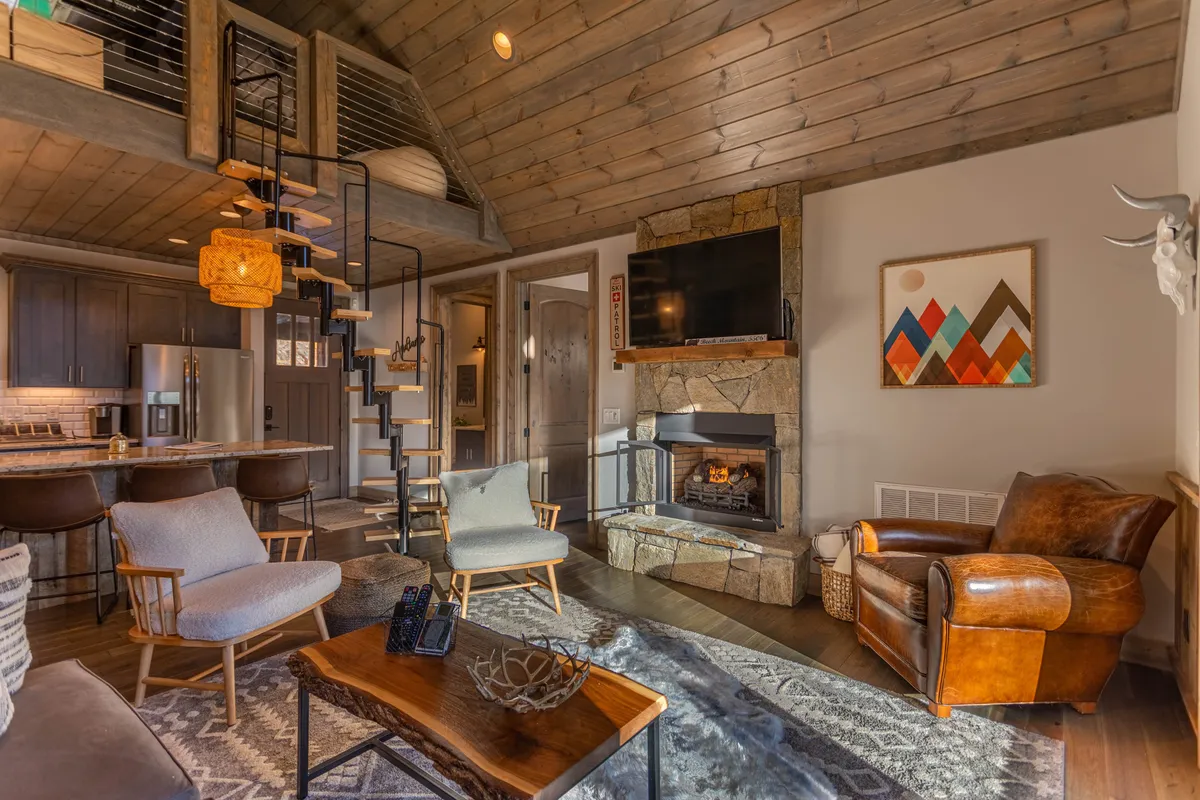 A welcoming open-concept living area featuring a cozy fireplace as the centerpiece, surrounded by comfortable seating options. The wooden ceiling and staircase add a rustic charm, while art and decor pieces encourage conversation. The space is designed for people to gather, relax, and socialize, evoking a sense of togetherness.