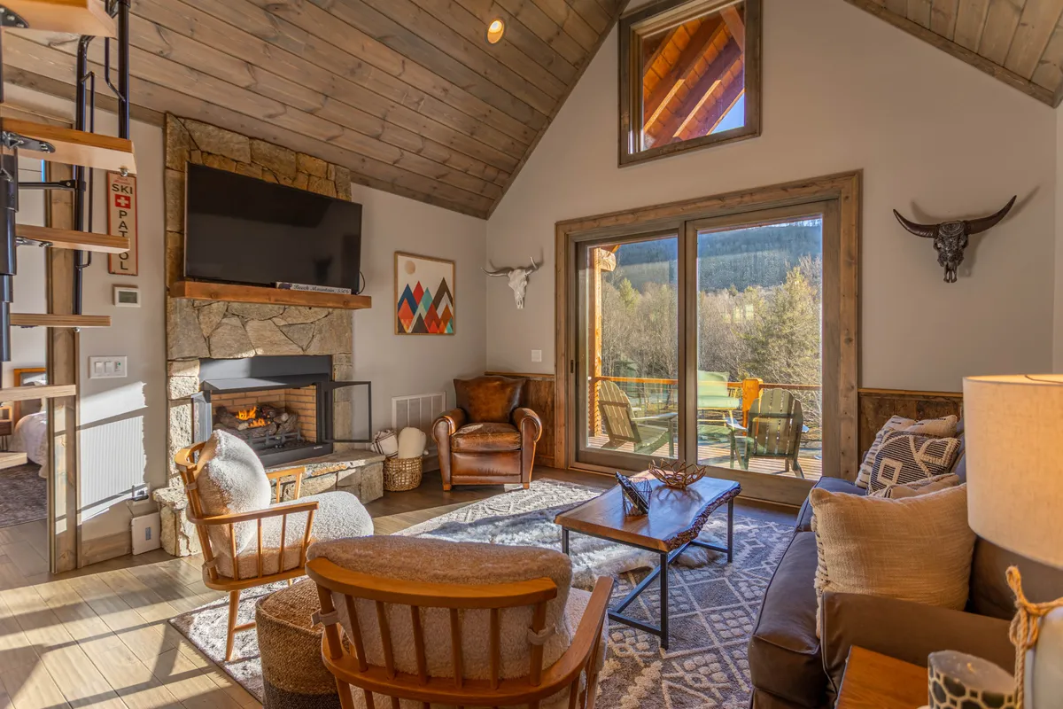 A cozy great room scene with a gas log fireplace as the focal point, surrounded by plush, inviting seating. Large windows offer natural light and a view outside, while the wooden ceiling adds warmth and character to the space.