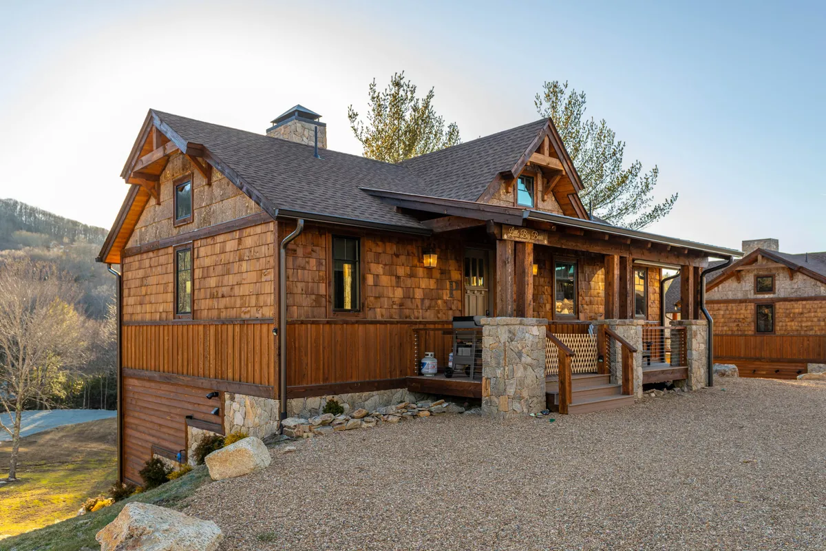A warm, inviting exterior shot featuring a wooden cabin with stone accents, a spacious porch, and lush landscaping. The scene hints at opportunities for outdoor relaxation and gathering, with the home nestled peacefully amidst greenery.