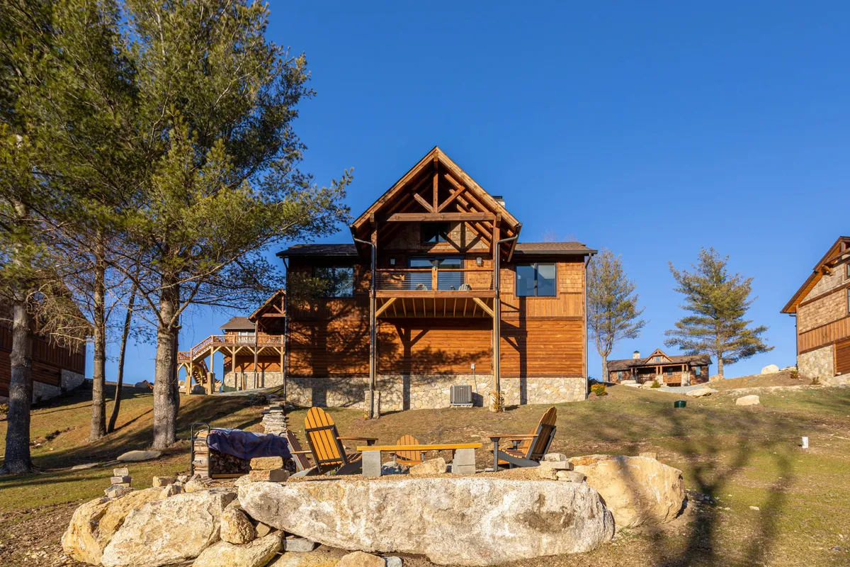 The image shows a charming wooden cabin nestled among tall trees, with a stone seating area and classic Adirondack chairs arranged around a fire pit. The setting is warm and inviting, suggesting opportunities for outdoor gatherings, stargazing, and soaking in the tranquil mountain atmosphere. Sunsets can be viewed from the deck, surrounded by the natural beauty of Banner Elk.