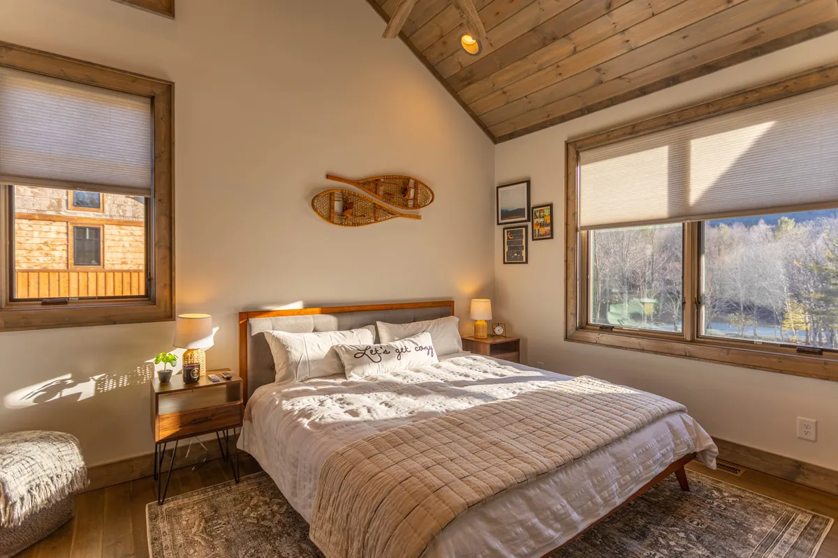 A peaceful bedroom featuring a king bed with decorative pillows, large windows overlooking nature, wood wall accents, and nightstands with lamps. The mood is calm and restful, ideal for unwinding after a long day.