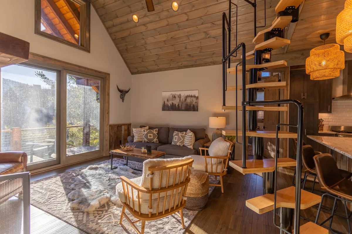 An interior view highlighting a spiral staircase leading up to a loft area, with large windows providing ample natural light. The space includes comfortable seating and decorative wall art, conveying a warm, welcoming mood ideal for relaxing or family gatherings.