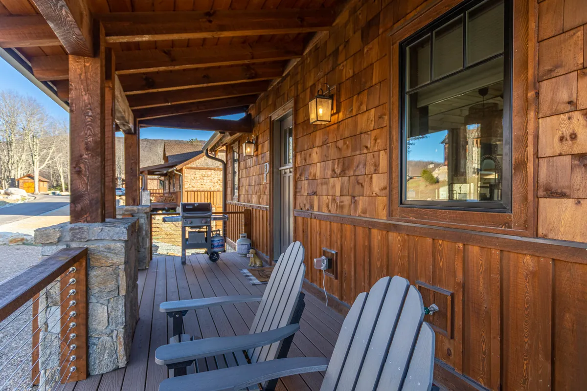 A close-up of a cozy outdoor seating area, showing Adirondack chairs positioned beside wooden siding and rock pillars, with a lantern light fixture adding warmth and charm. The scene feels intimate and welcoming, ideal for small gatherings or peaceful moments in nature.