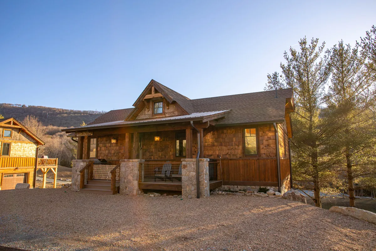 A cozy wooden porch with a stone facade is surrounded by tall trees and offers a sweeping mountain view. The inviting outdoor seating area looks perfect for relaxing, soaking in the tranquil setting, or enjoying a peaceful morning coffee while wrapped in nature.